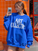 Person wearing a blue sweatshirt with 'ART COLLECTIVE' text, taking a mirror selfie against an orange tiled wall.